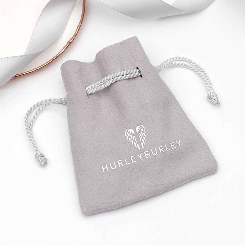 May include: A gray velvet pouch with a drawstring closure. The pouch has a white logo with a pair of angel wings and the text "HURLEYBURLEY".