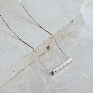 May include: A silver-toned necklace featuring a thin box chain and a horizontal rectangular bar pendant. The bar pendant has the name BRINLY engraved in a clean, sans-serif font on its front surface, creating a simple and polished appearance.