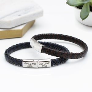May include: Two braided leather bracelets with a silver clasp. The bracelet on top is brown and black, and the bracelet on the bottom is blue and black. The bottom bracelet has a silver plate with the text "JAMES AND SARAH" and "1ST JUNE 2018".