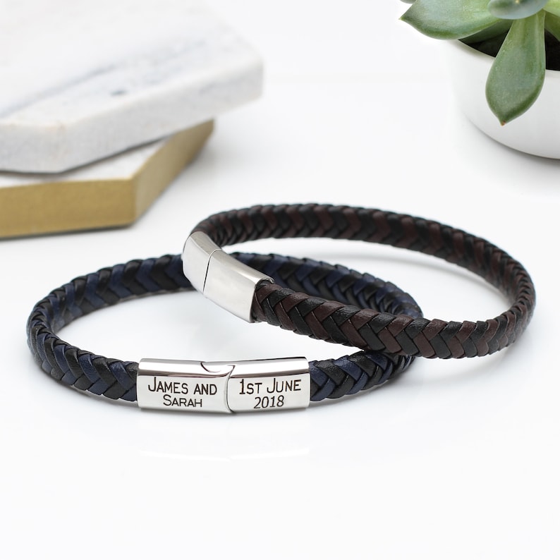 May include: Two braided leather bracelets with a silver clasp. The bracelet on top is brown and black, and the bracelet on the bottom is blue and black. The bottom bracelet has a silver plate with the text "JAMES AND SARAH" and "1ST JUNE 2018".