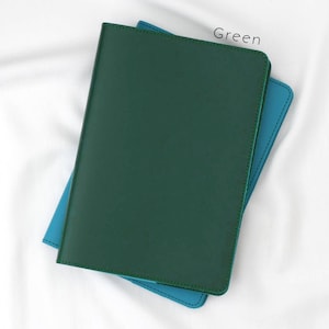 Puede incluir: Una cubierta de cuaderno de cuero verde con una cubierta de cuero azul debajo. El texto "Green" est&aacute; impreso en la esquina superior derecha de la imagen.