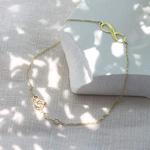 Könnte beinhalten: Ein goldfarbenes Armband mit einem Unendlichkeits-Anhänger und einer zarten Kette. Das Armband liegt auf einer weißen Oberfläche mit einem hellbraunen Stoffhintergrund.