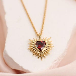 May include: Gold necklace with a heart-shaped, deep red gemstone pendant. The pendant has a sunburst design. The necklace is shown on a white surface with a soft pink background.