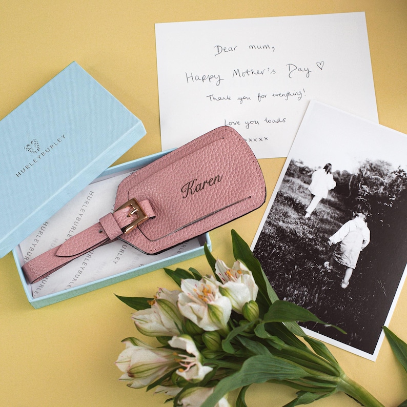 May include: A pink leather luggage tag with the name "Karen" embossed on it, presented in a light blue box. A handwritten card reads "Dear mum, Happy Mother's Day." A black and white photo and flowers complete the gift set.