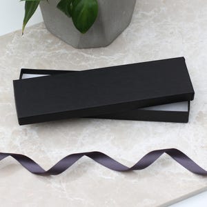 May include: An open black rectangular gift box with a white interior sits on a marble surface. A dark gray ribbon is in the foreground. A potted plant is in the background.
