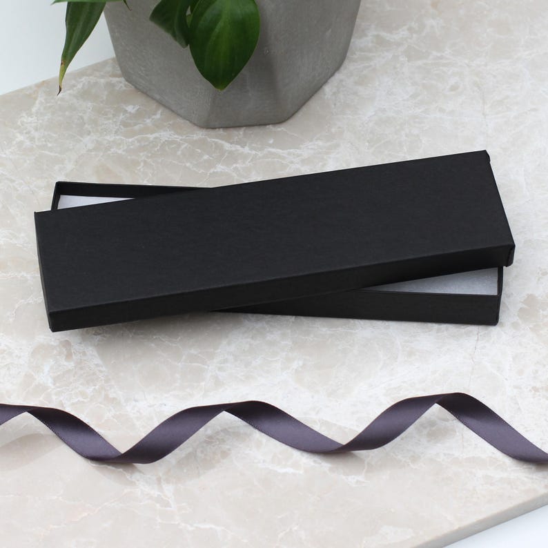 May include: An open black rectangular gift box with a white interior sits on a marble surface. A dark gray ribbon is in the foreground. A potted plant is in the background.