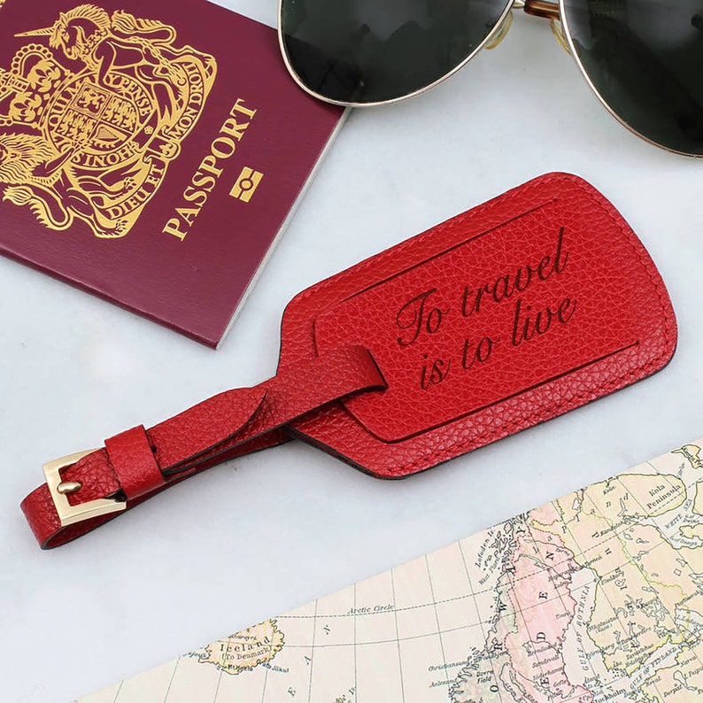May include: A red leather luggage tag with the text "To travel is to live" is displayed next to a passport and sunglasses. The tag has a gold-colored buckle and strap. A map is visible in the background.