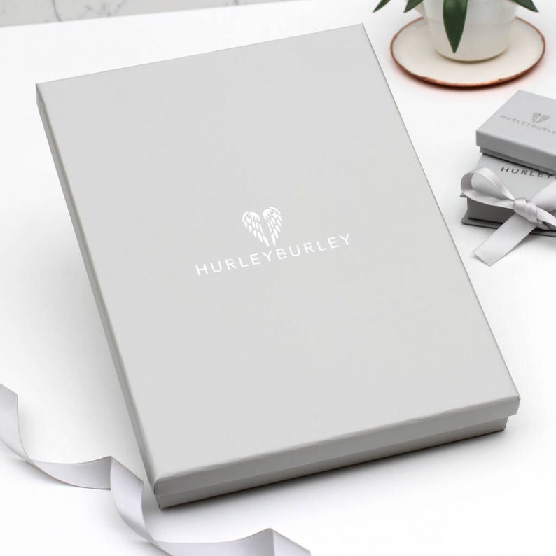 Puede incluir: Una caja de regalo gris con un logotipo blanco que dice "HURLEYBURLEY" y un par de alas de &aacute;ngel blancas.