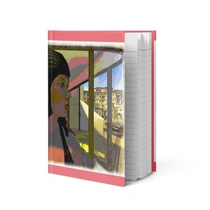 May include: A hardcover notebook with a pink border and a colorful illustration on the cover. The illustration depicts a person looking out a window at a cityscape. The notebook has lined pages.