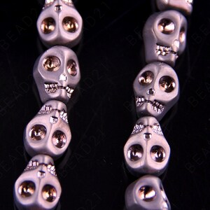 May include: A strand of gray skull beads with gold accents. The beads are strung on a black cord and have a detailed skull design.