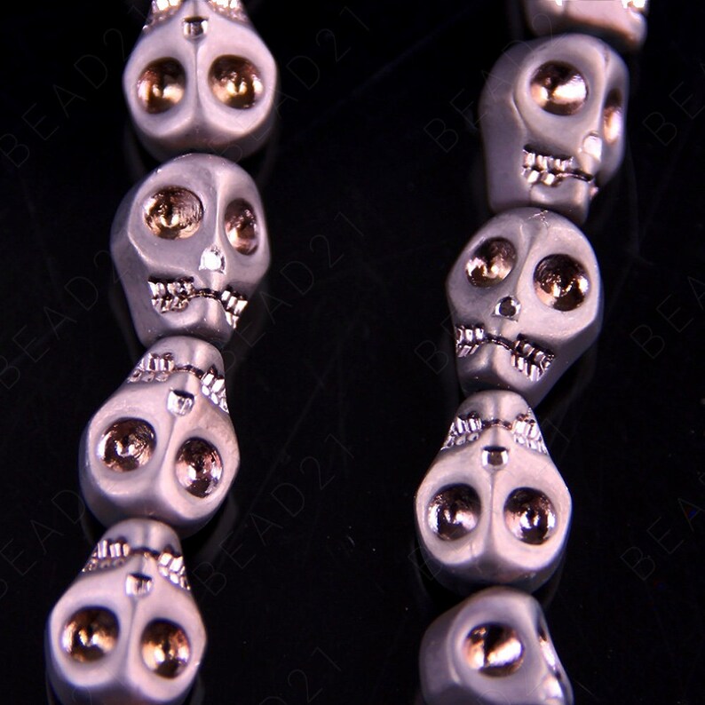 May include: A strand of gray skull beads with gold accents. The beads are strung on a black cord and have a detailed skull design.