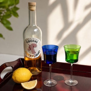 May include: Aperitivo Cocchi Americano bottle with a cork stopper, two stemmed glasses, one blue and one green, and a lemon on a wooden tray. The bottle has a label with text and a red and white design.