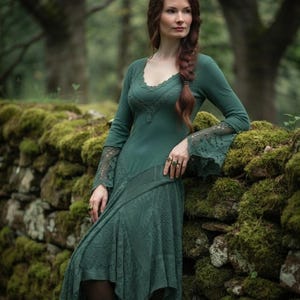 May include: A long-sleeved forest green dress featuring a scalloped neckline, intricate lace detailing on the flared cuffs, and a textured, asymmetrical skirt with layered fabric panels that fall below the knee in a pointed hemline.