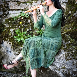 以下が含まれることがあります： 緑のレースのドレスを着た女性が、苔むした岩に座って木製の笛を吹いています。