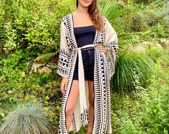 Puede incluir: Un kimono largo color crema con un patrón geométrico negro. El kimono está abierto por delante y atado a la cintura con un cinturón a juego. La persona lleva un top y unos pantalones cortos negros, y sandalias marrones.