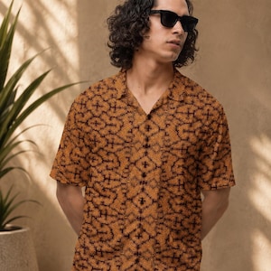 May include: A short-sleeved button-down shirt with a repeating pattern in shades of brown and black. The shirt is paired with blue pants. The person in the image is wearing black sunglasses.