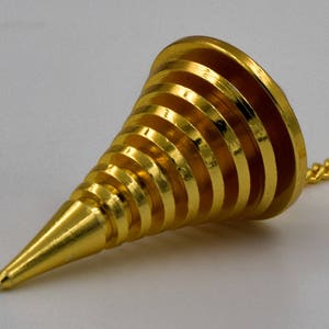 Può includere: Un filo a piombo in metallo color oro di forma conica, caratterizzato da una serie di creste orizzontali a gradini che si restringono verso una punta. L'oggetto è fissato a una sottile catena di metallo dorato che parte dall'estremità superiore.