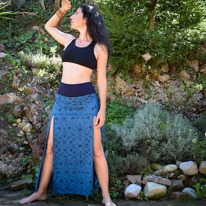 Può includere: Una donna indossa un reggiseno sportivo nero e una lunga gonna blu con un motivo geometrico e spacchi laterali. La gonna ha una cintura nera. È in piedi davanti a un muro roccioso con piante verdi.