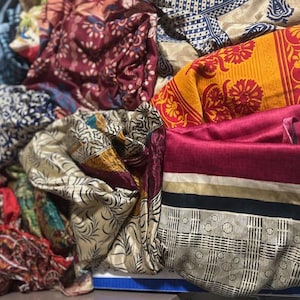 May include: A collection of colorful, patterned fabrics. The textiles display a variety of designs, including floral, geometric, and abstract patterns. Colors include red, yellow, blue, and cream. The fabrics appear to be silk or a similar lightweight material.