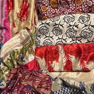 May include: A collection of colorful fabrics with various patterns. The fabrics include floral, paisley, and geometric designs in shades of red, cream, burgundy, and blue. Some fabrics have a silky sheen, while others appear more matte.