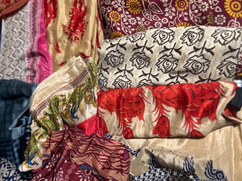 May include: A collection of colorful fabrics with various patterns. The fabrics include floral, paisley, and geometric designs in shades of red, cream, burgundy, and blue. Some fabrics have a silky sheen, while others appear more matte.
