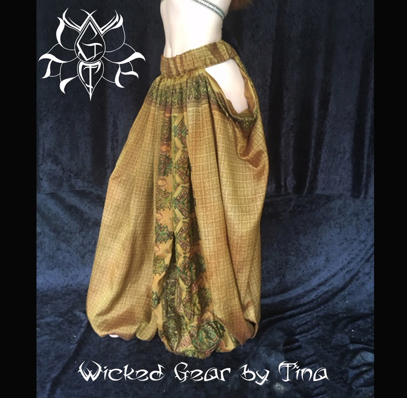 May include: A pair of flowing, patterned pants in shades of gold and olive green. The pants feature a unique design with cutouts and a central panel with intricate floral and paisley motifs. The text "Wicked Gear by Tina" is visible at the bottom.
