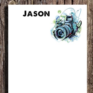 May include: A white notepad with the name "JASON" printed in black at the top. A colorful illustration of a camera with a blue lens is in the upper right corner.