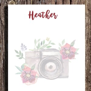 May include: A watercolor illustration of a vintage camera with pink and purple flowers and green leaves. The name "Heather" is written in red at the top of the image.