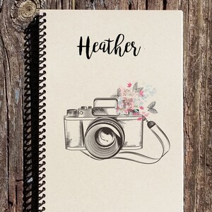 May include: A cream-colored spiral bound notebook with the name "Heather" printed in black cursive. The cover features a black and white illustration of a vintage camera with pink and blue flowers.