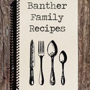 Puede incluir: Un cuaderno marr&oacute;n de espiral con el texto "Banther Family Recipes" impreso en negro. La portada presenta una ilustraci&oacute;n en blanco y negro de un cuchillo, un tenedor y dos cucharas.