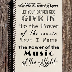 May include: A notebook with a spiral binding and a quote about music. The quote reads: "Let the Dream Begin Let Your Darker Side Give In To the Power of the Music That I Write The Power of the Music of the Night"