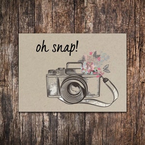 May include: A vintage camera with a floral design on top. The camera is brown and black with a white strap. The text "oh snap!" is written above the camera.