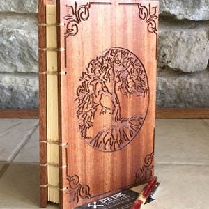 May include: A wooden journal with a tree of life design carved into the front cover. The journal is bound with a leather spine and has a cream-colored paper interior. A business card for "8th Line Creations" is in the foreground.