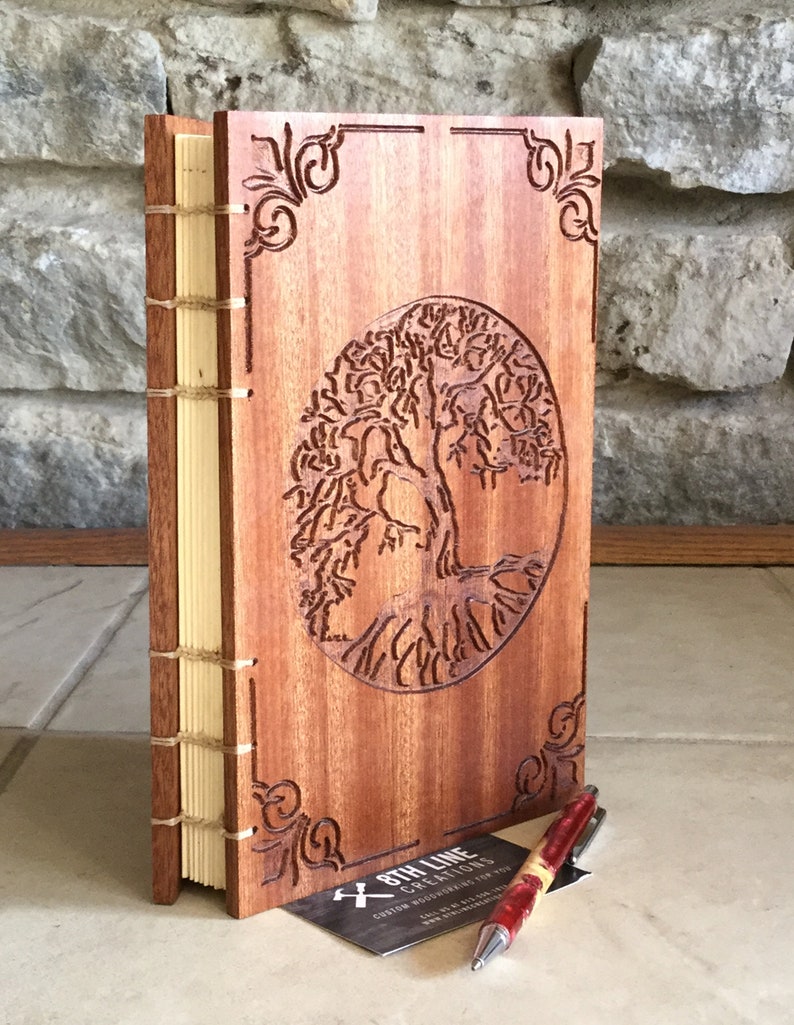 May include: A wooden journal with a tree of life design carved into the front cover. The journal is bound with a leather spine and has a cream-colored paper interior. A business card for "8th Line Creations" is in the foreground.