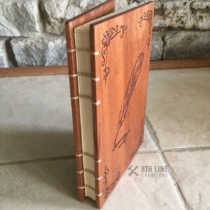 May include: A wooden journal with a carved feather and quill design. The journal is bound with a cream-colored spine and has a brown wood cover. The text "8TH LINE CREATIONS" is visible on the bottom right corner.