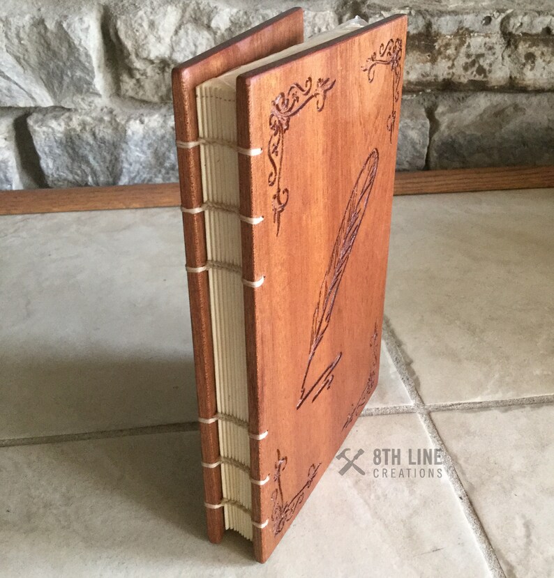 May include: A wooden journal with a carved feather and quill design. The journal is bound with a cream-colored spine and has a brown wood cover. The text "8TH LINE CREATIONS" is visible on the bottom right corner.