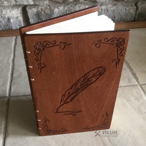 May include: A brown wooden journal with a feather and ink design engraved on the cover. The journal is closed and has a white spine. The text "8TH LINE CREATIONS" is visible on the bottom right corner of the cover.