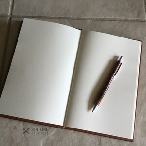 May include: An open brown leather bound journal with blank white pages and a silver pen with a brown and red marbled barrel lying on the open pages. The journal has the text "8th Line Creations" printed on the bottom left corner.