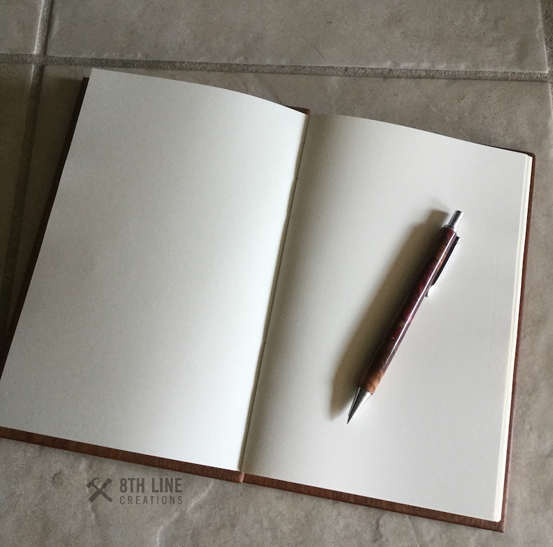 May include: An open brown leather bound journal with blank white pages and a silver pen with a brown and red marbled barrel lying on the open pages. The journal has the text "8th Line Creations" printed on the bottom left corner.
