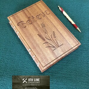 May include: A wooden journal with the word "Believe" carved into the cover. The journal also features a design of wheat stalks. The journal is sitting on a green surface with a red and white pen next to it.