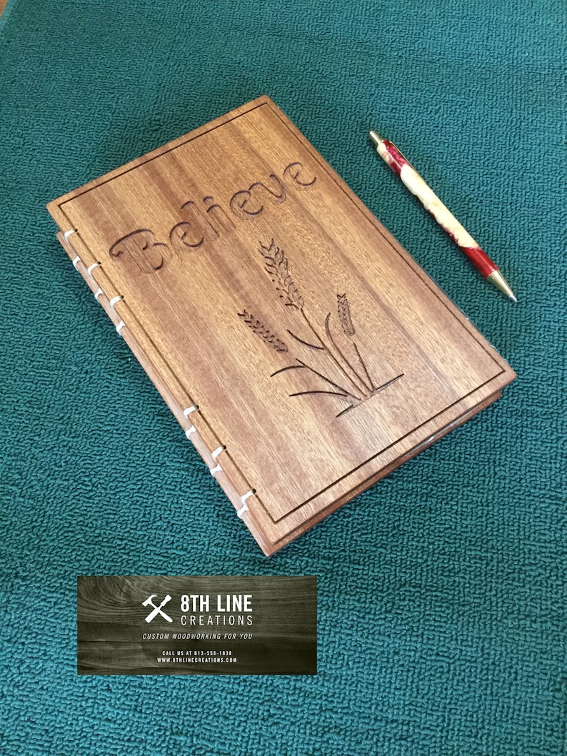 May include: A wooden journal with the word "Believe" carved into the cover. The journal also features a design of wheat stalks. The journal is sitting on a green surface with a red and white pen next to it.
