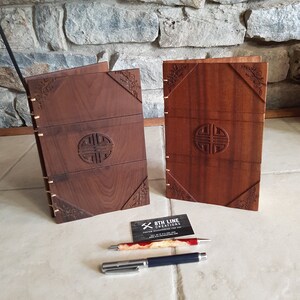 May include: Two wooden journals with intricate carvings. The covers are a rich brown color and feature a circular design with a knotwork pattern. The journals are closed and standing upright on a tiled surface.