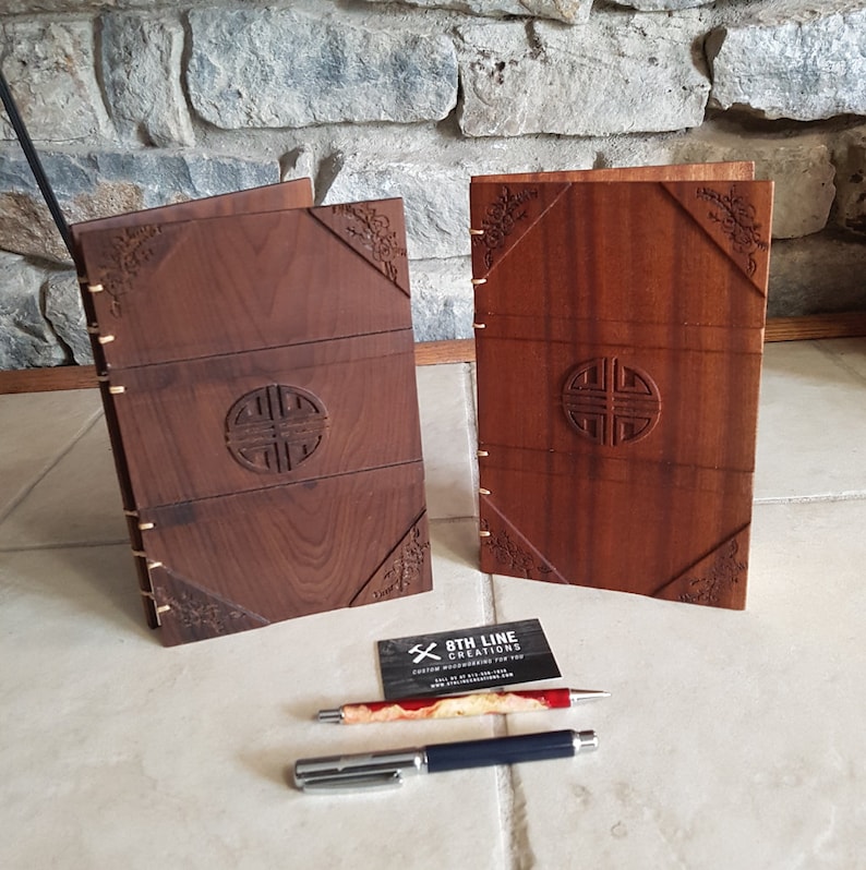 May include: Two wooden journals with intricate carvings. The covers are a rich brown color and feature a circular design with a knotwork pattern. The journals are closed and standing upright on a tiled surface.