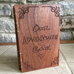 May include: A wooden journal with a brown wood cover and the words "Our Adventure Book" carved into the front. The cover has decorative carvings around the edges.