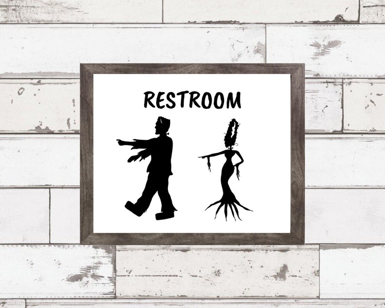 May include: A black and white sign with the word "RESTROOM" in a handwritten font. The sign features a silhouette of Frankenstein's monster and a female monster with long hair and a tree-like body.