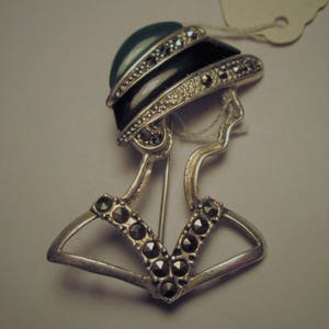 May include: A vintage silver-toned brooch in the shape of a woman's profile. The brooch features a black and green hat with marcasite accents and a V-shaped collar adorned with dark stones. A tag is attached.
