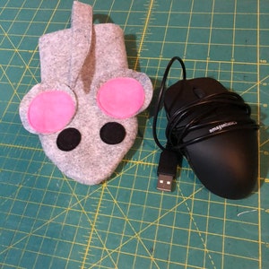 May include: A gray felt mouse-shaped computer mouse holder with pink ears and black eyes. A black computer mouse with a USB connector and the text "amazonbasics" is on a green cutting mat.