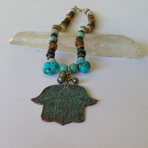 Op de afbeelding: Een turquoise en bruine kralenketting met een grote turquoise hamsa hand hanger. De ketting ligt op een witte achtergrond.