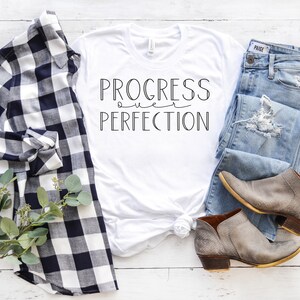 May include: White t-shirt with the text "PROGRESS over PERFECTION" in black lettering. The shirt is styled with a plaid shirt, jeans, and ankle boots.