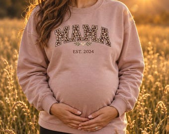 May include: A dusty rose long-sleeve sweatshirt featuring the word MAMA in a leopard print pattern with a black outline. Below the text is a small green leaf graphic and the text EST. 2024 printed in a simple black font.